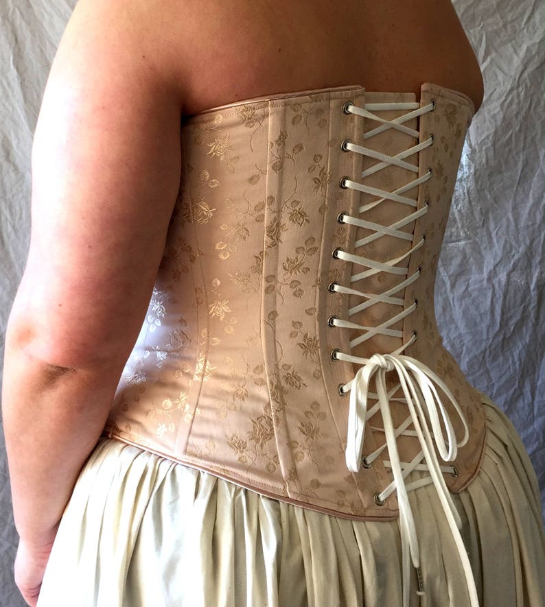 May include: A beige corset featuring a subtle floral jacquard pattern, constructed with vertical panels and silver-toned metal grommets. The back is secured with a white ribbon lace-up closure tied in a bow at the bottom, worn over a cream skirt.