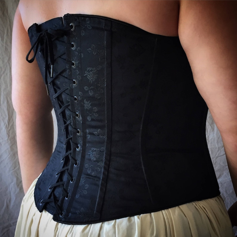 May include: A black corset featuring a subtle tonal floral jacquard pattern, a straight neckline, and a back closure with black lacing threaded through metal grommets. The garment is worn over a light-colored skirt, showcasing the structured design.