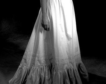 May include: A white cotton petticoat with a ruffled hem and three rows of horizontal stitching. The petticoat is worn by a person standing on a dark background.