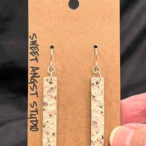 May include: A pair of dangle earrings with a beige and brown speckled rectangular design. The earrings are hanging on a brown card with the text "Sweet Angst Studio".