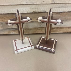 May include: Two wooden crosses with layered designs. One cross has a light wood base with a white and brown cross design, while the other has a dark wood base with a similar design. The crosses are made of various wood tones.