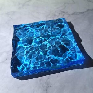 May include: A square, transparent resin piece with a blue water effect. The surface mimics the look of water with light reflecting off the surface. The edges are a darker blue, creating a contrast. The piece is sitting on a light grey surface.