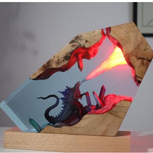 May include: A resin and wood sculpture of a monster breathing fire, set on a wooden base. The monster is blue and purple, and the fire is orange and yellow. The sculpture is lit from within, creating a glowing effect.