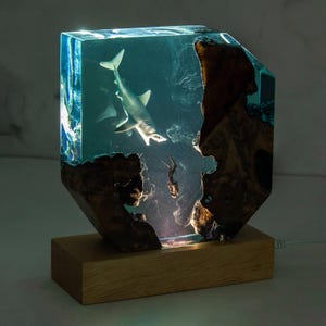 Peut inclure: Une sculpture en résine d'un requin nageant dans un bloc de résine bleu transparent. Le bloc est monté sur une base en bois. Le requin est blanc et a un œil noir. Le bloc de résine est éclairé par derrière, créant un effet lumineux.