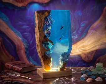 May include: A vertical lamp featuring a natural wood edge combined with translucent blue resin. The resin section contains miniature scuba diver figures and rock formations. The base is a rectangular wood block with internal lighting.