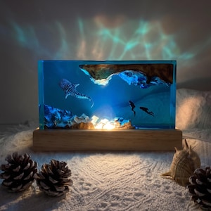 Peut inclure: Une lampe rectangulaire en résine représentant une scène sous-marine bleu profond avec un requin-baleine, deux plongeurs et des éléments en bois naturel. L'objet est monté sur un socle en bois clair et émet une lumière douce créant des reflets.