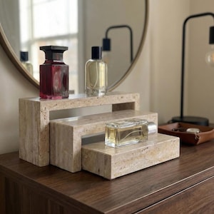 May include: Three perfume bottles on a tiered, rectangular stone display. The bottles are various shapes and colours, including a dark red, clear, and light yellow. The display is made of a light beige stone and sits on a dark brown wooden surface.