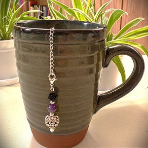 May include: A dark green ceramic mug with a ribbed texture and a brown base. A silver-toned metal chain hangs from the rim, featuring a black rose bead, a round purple stone bead, and a silver-toned metal heart charm with intricate cutouts.