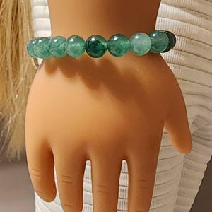 May include: A green beaded bracelet. The bracelet features round, polished beads in varying shades of green. The beads are strung together, forming a complete circle. The bracelet is worn on a wrist.