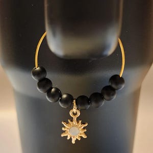 May include: A gold-colored hoop earring with a strand of black beads and a sun-shaped charm. The sun charm has a gold outline and a white center. The earring is hanging on a black surface.