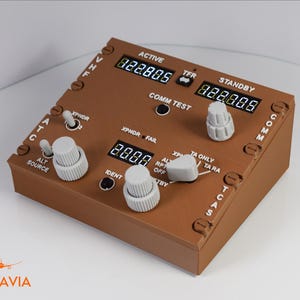 May include: A brown aviation radio control panel featuring digital displays for active and standby frequencies, labelled VHF, COMM, ATC, and TCAS. The device includes multiple grey rotary knobs, toggle switches, and push buttons with white text labels.