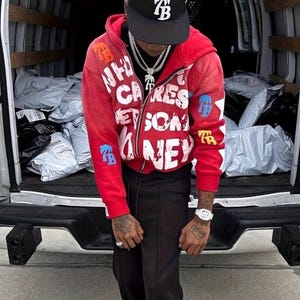 Op de afbeelding: Een rode zip-up hoodie met witte tekst en blauwe en gouden accenten. De hoodie heeft de tekst "WHO CARES GET SOME MONEY" en de letters "TB" op de mouwen. De persoon draagt een zwarte baseball cap met de letters "TB".