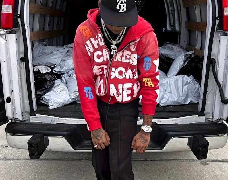 Op de afbeelding: Een rode zip-up hoodie met witte tekst en blauwe en gouden accenten. De hoodie heeft de tekst "WHO CARES GET SOME MONEY" en de letters "TB" op de mouwen. De persoon draagt een zwarte baseball cap met de letters "TB".