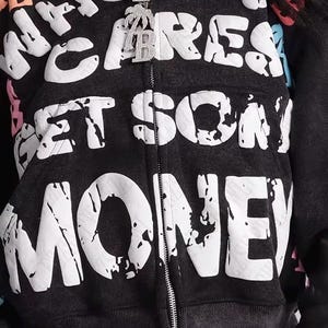 Op de afbeelding: Een donkergrijze hoodie met ritssluiting met de woorden "WHO CARES GET SOME MONEY" in grote witte letters. De hoodie heeft een distressed look en een zilveren rits. Een zilveren hanger met een palmboom en de letter "B" hangt aan de rits.