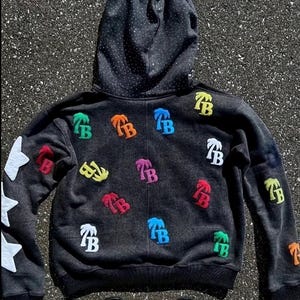 Op de afbeelding: Een donkergrijze hoodie met een zwarte gestippelde capuchonvoering en witte steraccenten op de linkermouw. De hoodie heeft kleurrijke geborduurde palmboom- en "TB"-logopatches.