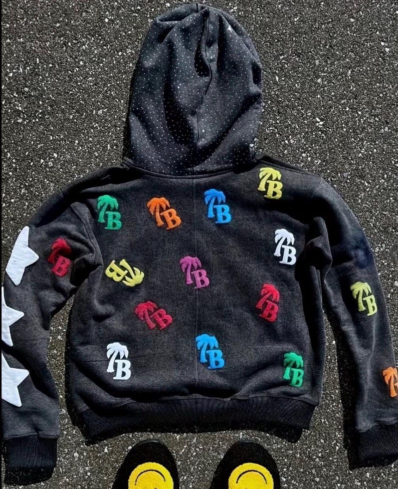 Op de afbeelding: Een donkergrijze hoodie met een zwarte gestippelde capuchonvoering en witte steraccenten op de linkermouw. De hoodie heeft kleurrijke geborduurde palmboom- en "TB"-logopatches.