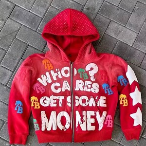 Op de afbeelding: Rode hoodie met ritssluiting met de tekst "WHO CARES GET SOME MONEY" in het wit. De hoodie heeft witte sterontwerpen op de linkermouw en kleurrijke patches. De capuchon is bedekt met kleine, witte stippen.