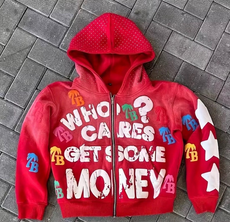 Op de afbeelding: Rode hoodie met ritssluiting met de tekst "WHO CARES GET SOME MONEY" in het wit. De hoodie heeft witte sterontwerpen op de linkermouw en kleurrijke patches. De capuchon is bedekt met kleine, witte stippen.