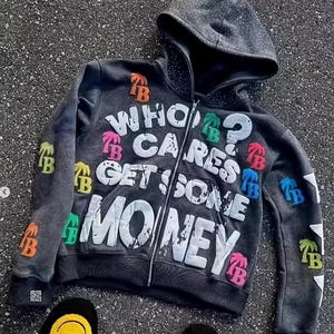 Op de afbeelding: Een donkergrijze hoodie met ritssluiting met de tekst "WHO CARES GET SOME MONEY" in het wit. De hoodie heeft kleurrijke patches en een gestippelde capuchon. Zwarte slippers met smiley-gezichten zijn onderaan zichtbaar.