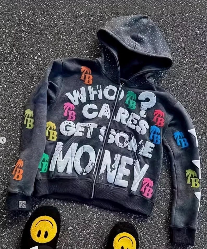 Op de afbeelding: Een donkergrijze hoodie met ritssluiting met de tekst "WHO CARES GET SOME MONEY" in het wit. De hoodie heeft kleurrijke patches en een gestippelde capuchon. Zwarte slippers met smiley-gezichten zijn onderaan zichtbaar.