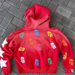 Op de afbeelding: Rode hoodie met een gradi&euml;nteffect, met witte steraccenten op de linkermouw en kleurrijke palmboom- en "TB"-logopatches. De capuchon is gestippeld met kleine witte accenten.