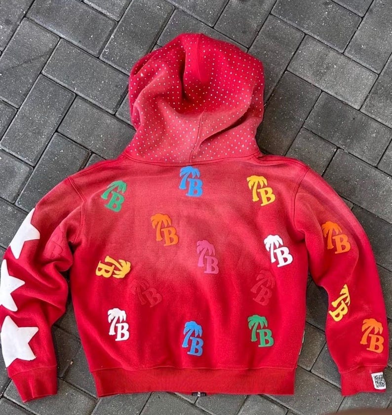 Op de afbeelding: Rode hoodie met een gradi&euml;nteffect, met witte steraccenten op de linkermouw en kleurrijke palmboom- en "TB"-logopatches. De capuchon is gestippeld met kleine witte accenten.