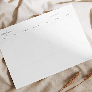 Könnte beinhalten: Ein weißer Wochenplaner mit dem Wort "Wochenplaner" in Schreibschrift. Der Planer hat Abschnitte für jeden Wochentag, wobei die Wochentage auf Deutsch geschrieben sind. Der Planer liegt auf einem beigen Stoff.