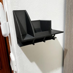 Op de afbeelding: Een zwarte wandorganizer met meerdere compartimenten en haken. De organizer heeft een plank, een gedeelte voor het bewaren van spullen en drie haken om spullen op te hangen. Het is aan een witte muur bevestigd.