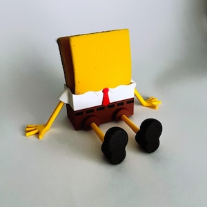 Könnte beinhalten: Eine gelbe Schwammkreatur mit braunen Hosen, einem weißen Hemd und einer roten Krawatte. Die Figur sitzt mit ausgestreckten Armen und Beinen. Der Schwamm besteht aus einem weichen, saugfähigen Material.