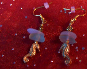 May include: A pair of gold tone earrings with a seahorse charm and a light blue glass bead. The earrings are on a red sparkly background.