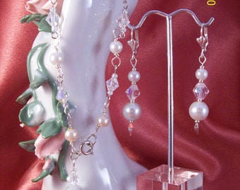 Puede incluir: Un conjunto de pulsera y pendientes de plata con perlas blancas y detalles de cristal. La pulsera está en una mano de porcelana blanca y los pendientes están en un soporte de acrílico transparente.