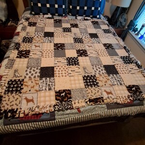 Op de afbeelding: Een patchwork deken met honden thema, met vierkanten in beige, bruin en zwart. De deken heeft honden silhouetten, pootafdrukken en bot patronen. De randen zijn afgewerkt met een gefranjerde rand.