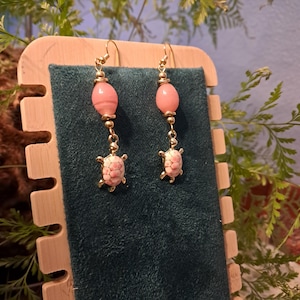 Może przedstawiać: Złote kolczyki z różowymi zawieszkami w kształcie żółwi i owalnymi różowymi koralikami. Kolczyki mają delikatny design z małymi złotymi koralikami i haczykami. Żółwie mają szczegółowy wzór skorupy.