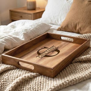 May include: A rectangular wooden serving tray with cut-out handles. The tray is engraved with the name "Giullano" in a stylized font. The wood has a warm, natural tone, and the tray is resting on a textured, beige blanket.