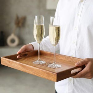 May include: A wooden serving tray with two champagne flutes filled with bubbly liquid. The tray has a rectangular shape with cut-out handles and a personalized inscription. The flutes are clear glass with long stems.