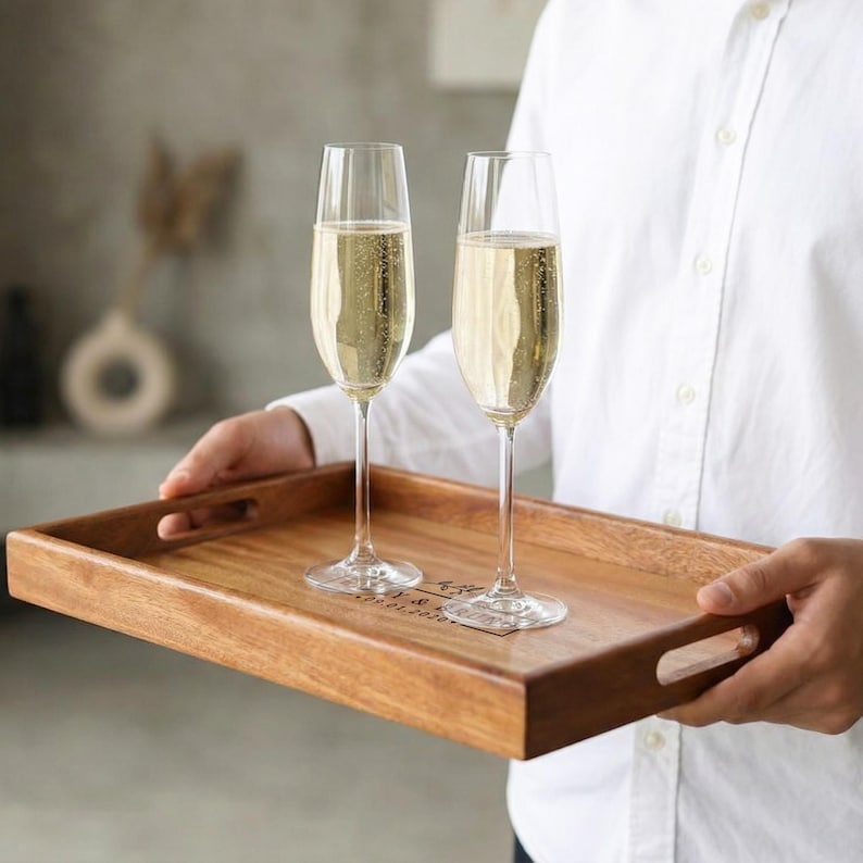 May include: A wooden serving tray with two champagne flutes filled with bubbly liquid. The tray has a rectangular shape with cut-out handles and a personalized inscription. The flutes are clear glass with long stems.