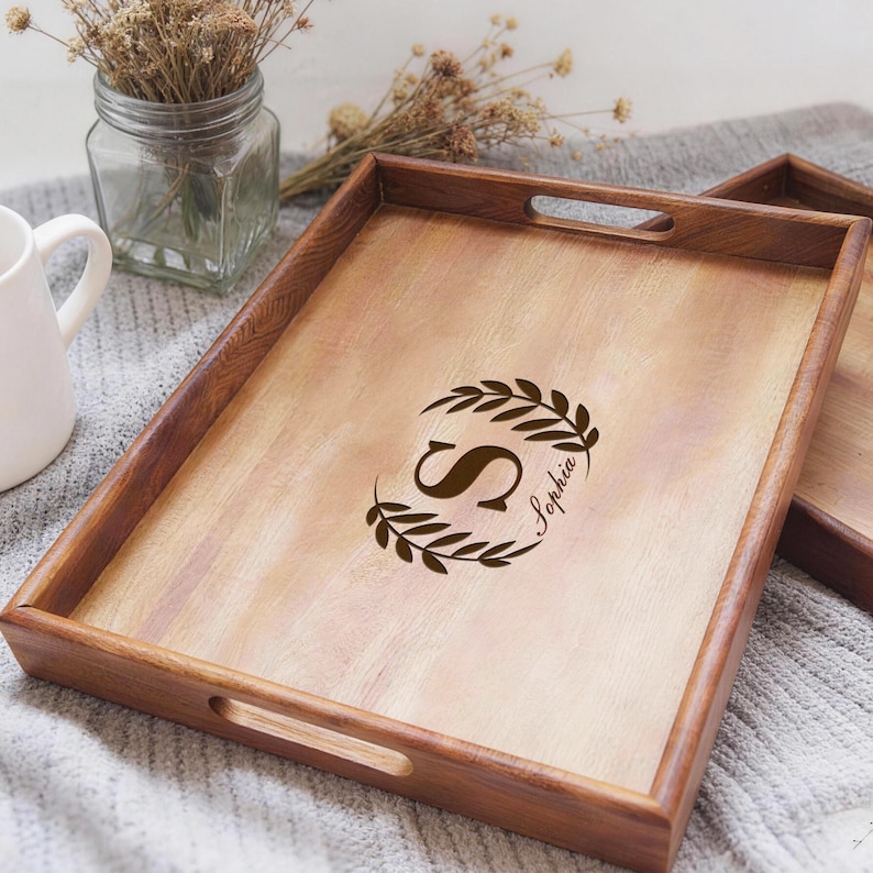 May include: A rectangular wooden serving tray with a dark brown frame and a light brown interior. The tray features a decorative design with the letter "S" and the name "Sophia" in a circular wreath. The tray has cut-out handles.