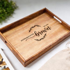 May include: A rectangular wooden serving tray with a dark brown frame and light brown interior. The tray features the names "Alyssa & Justin" and the last name "Brown" with the year "EST.2024" in a decorative script. The tray has two handles and is surrounded by a light-colored background.