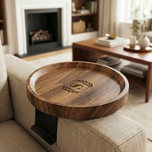 May include: A round wooden sofa tray with a dark finish, featuring a laser-etched monogram and name within a laurel wreath. The tray is supported by a black metal arm that rests on the sofa arm. The tray is designed for convenience and style.
