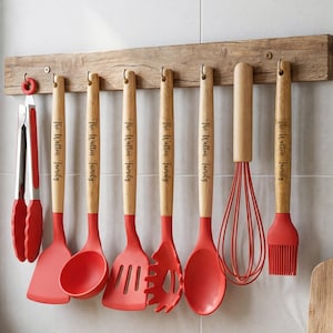 May include: A collection of red and wood-handled kitchen utensils hanging on a wooden rack. The set includes tongs, a spatula, a ladle, a slotted turner, a pasta spoon, a whisk, and a basting brush. Each handle is engraved with "The Walton Family."
