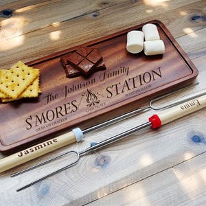 May include: A wooden s'mores serving board with the text "The Johnson Family S'mores Station" engraved. The board holds graham crackers, chocolate squares, and marshmallows. Two s'mores roasting sticks are included, one labeled "Jasmine" and the other "Emma".