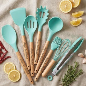 May include: A selection of teal silicone kitchen utensils with wooden handles. The set includes a spatula, slotted spoon, pasta server, ladle, basting brush, whisk, and tongs. Each handle is engraved with "Harper love Michael". Lemons, rosemary, and garlic are also present.
