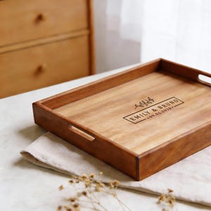 May include: A rectangular wooden serving tray with a natural wood grain finish. The tray has cut-out handles and features the engraved text "EMILY & BRUNO - 09.01.2020" with a floral design. The tray is resting on a beige cloth.