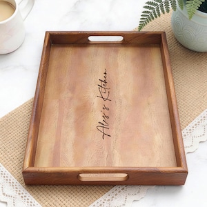 May include: A rectangular wooden serving tray with cut-out handles. The tray has a light brown interior and a darker brown frame. The words "Abby's Kitchen" are written in a cursive font on the tray's surface. The tray is on a beige surface.