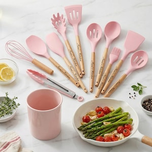 May include: A set of pink kitchen utensils with wooden handles, including spatulas, spoons, a whisk, and tongs. The handles are engraved with "Nana's Kitchen". A pink ceramic utensil holder and a pan of cooked vegetables are also present.