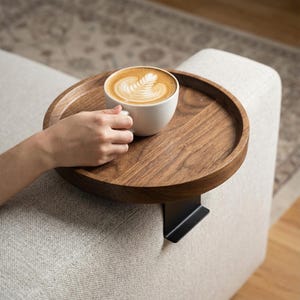 May include: A round, dark wood sofa arm tray with a black metal bracket, holding a white coffee cup filled with a latte. The latte art features a leaf design. The tray is attached to a light-colored sofa.