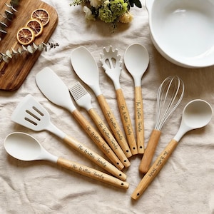 Può includere: Un set di utensili da cucina in silicone bianco con manici in legno, tra cui una spatola, un cucchiaio, una frusta e un pennello. I manici sono incisi con "The Walton Family".
