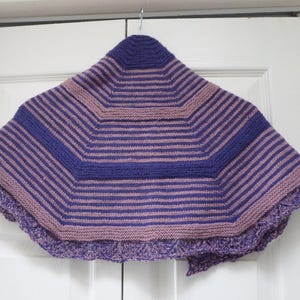 May include: A handmade, knitted shawl in shades of purple and mauve, with a striped pattern. The shawl has a unique geometric shape and a decorative, scalloped edge. The item is hanging on a white door.