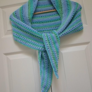 May include: A hand-knitted shawl in shades of blue and green, hanging on a wooden hanger. The shawl features a striped pattern with a delicate openwork design. The ends are tied in a knot, showcasing the intricate craftsmanship.