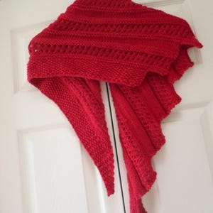 May include: A red knitted shawl with a lacy pattern. The shawl is hanging on a white door.